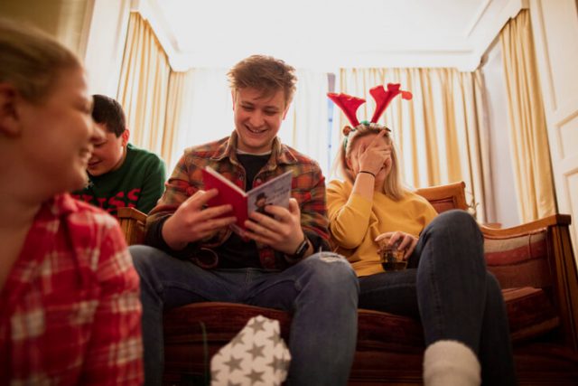Young people are sitting together in the living room at a house party, opening Christmas presents.