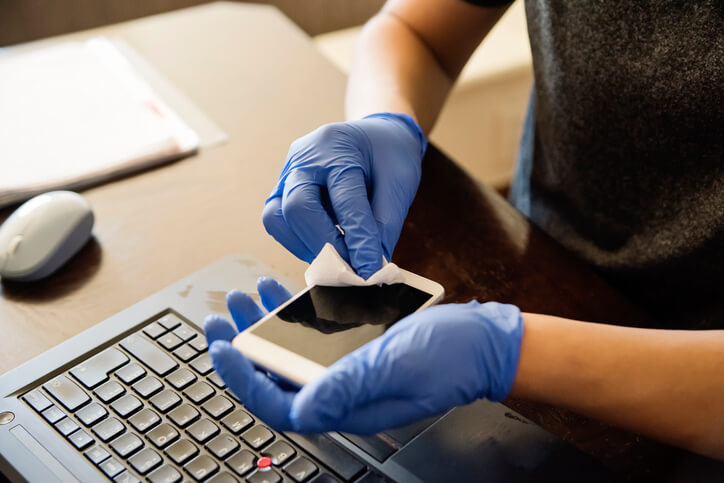 Man with gloves hands wiping mobile phone with disinfectant wipe while working from home. Horizontal indoors close-up with copy space.