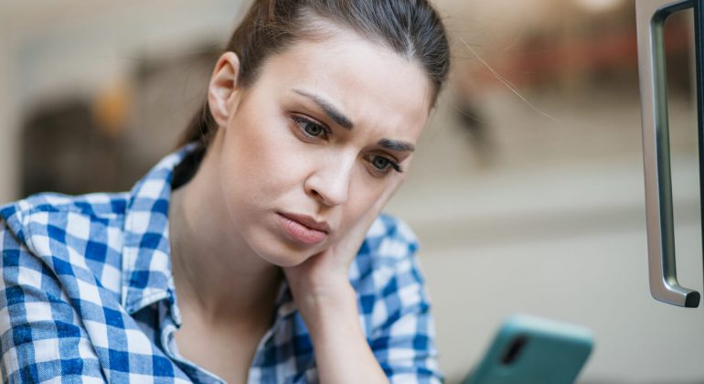 Close up of a depressed Caucasian young woman sitting at home, reading some bad news on her smart phone, feeling sad and worried